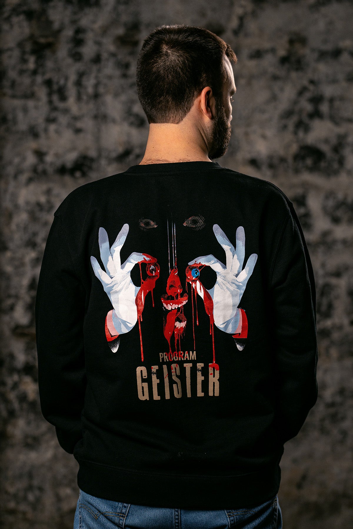 A man viewed from behind wearing a black crewneck sweatshirt with a large graphic print on the back. The graphic features a dripping red skull, white gloved hands forming an 'OK' sign, and the text 'PROGRAM GEISTER'.